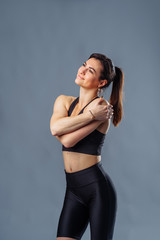 Pretty Hispanic personal trainer and diet coach standing isolated on gray background. Health and fitness concept - portrait of an caucasian american girl posing with fitness clothes. Hugging herself