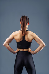 Fototapeta na wymiar Pumped up strong fitness woman showing back and biceps muscles strength. Fit girl fitness model isolated on gray background. Hands up, hands on the waist line. Trainer wearing black sportwear