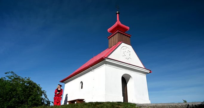Sad woman lady in red historic victorian dress with hat walking from church. Little white chapel.