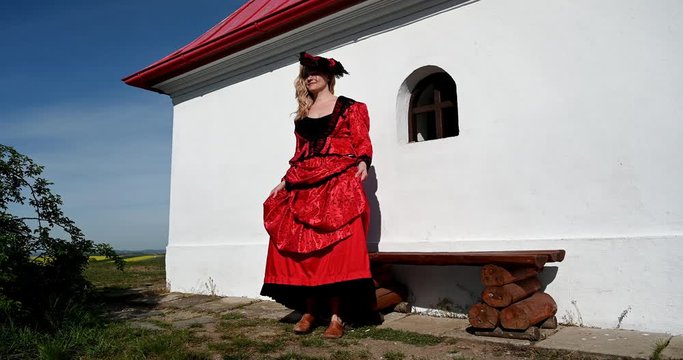 Blond lady in red costume dress sitting in nature near chapel relaxing