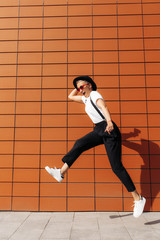Fototapeta premium Carefree hipster young woman wearing red sunglasses and black hat jumping near wall of modern building.