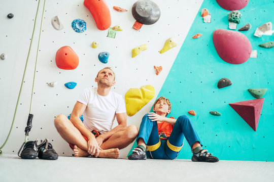 Father and teenage son sitting near the indoor climbing wall. They resting after the active climbing. Happy parenting concept image.