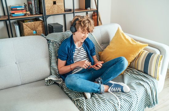 Preteen boy sitting at home on cozy sofa dressed casual jeans and new sneakers listening to music and chatting using wireless headphones connected with smartphone. Child use electronic devices concept