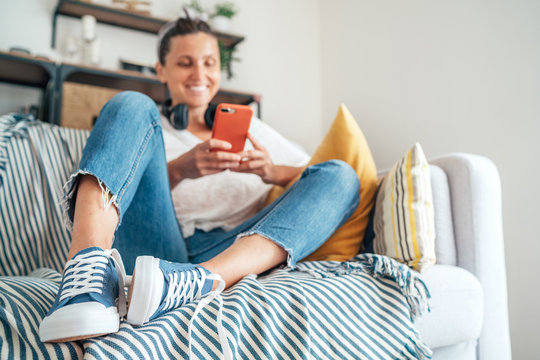 Young female sitting on cozy comfortable living room sofa, smiling and chatting with friends using modern smartphone. Selective focus on new blue sneakers. Comfortable clothing and footwear concept