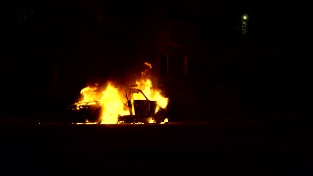 Car completely engulfed in fire near a residential building in the city. Theme of revenge through damage and destruction of property. Building close to burning automobile.