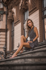 Beautiful blonde young girl wearing fashionable clothes sitting on the stairs in a blue sverfan bassek against the background of a light building