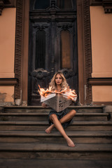 beautiful blonde girl in a sarofan, bassek sitting on the stairs and holding a burning newspaper against the background of the old building