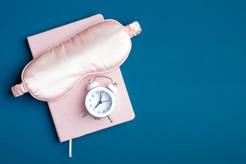 Pink sleep eye mask, dream book and alarm clock on dark blue background. Good night, insomnia...