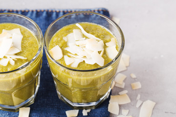 Green smoothies with coconut flakes in glass on denim napkin, gray background. Health content, healthy diet. Top view