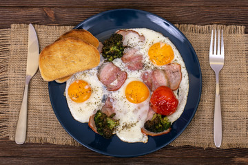 Delicious breakfast with fried eggs, bacon, tomato, broccoli and toasts in the plate that is on burlap, on wooden background, top view