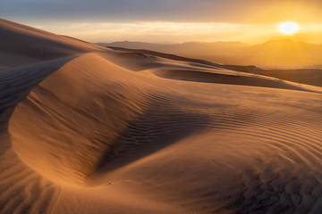 Obraz na płótnie Canvas The desert sand is blowing with the strong evening wind creating waves and patterns in the dune as the sun is setting under cloudy sky over the mountains in the distance.