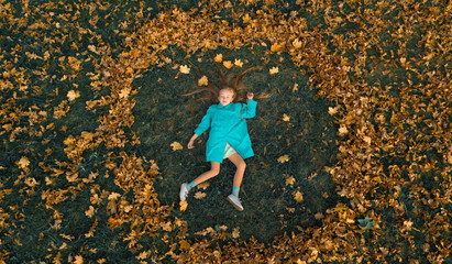 A little girl in a blue coat lies on the green grass and around her spread around in a circle of yellow leaves. Photo taken from the top of the drone