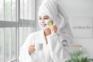 Beautiful young African-American woman with coffee and facial mask in bathroom