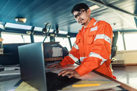 Filipino deck Officer on bridge of vessel or ship wearing coverall during navigaton watch at sea . He is using laptop, electronic paperwork at sea, concept of reporting