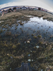 Feet with shoes and rain puddle and reflection on the ground with rural landscape and sunset with clouds and blue and purple tones