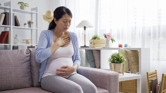 Portrait Of Young Asian Chinese Pregnant Woman Sitting On Couch Hands Touching Heart Feeling Nauseous In 1st Trimester Of Pregnancy. Copy Space. Morning Sickness In Maternal Time Concept.