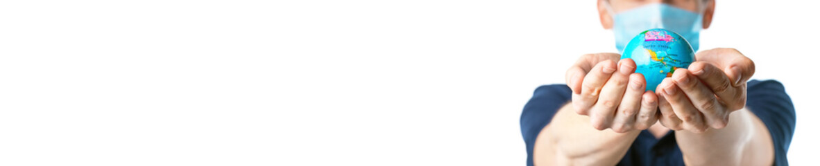  Close-up of a man's hands holding a small globe, with images USA, Canada, Bahamas other,  a man in a medical mask out of focus, dressed in a t-shirt, on a white  background. Coronavirus, Covid-19