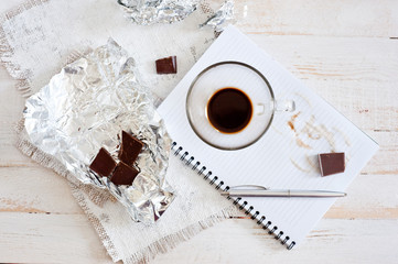 A cup of coffee, chocolate, a sheet of paper and a pen on a white wooden surface. Background for...