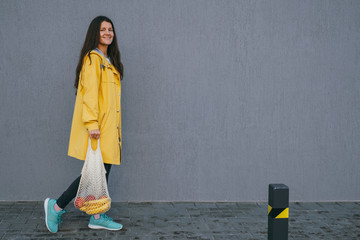 young woman in a yellow raincoat carrying string bag with fresh bananas and apples on the street