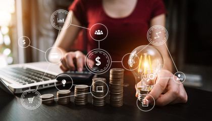 business hand holding lightbulb with using laptop computer and money stack in office. idea saving energy and accounting finance concept  in morning light
