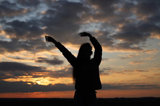 Silhouette of a young girl with hands up at sunset. Feels happy and raises a hand. Sunset time.
