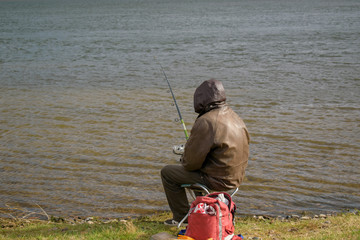 Fisherman is fishing. Man with a spin sits by a river or lake. View from the back, hood. Brown leather jacket. Bag with gear and fishing accessories. 