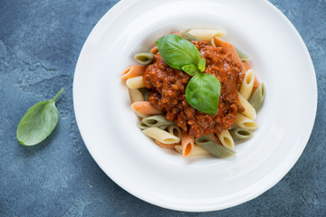 White plate with italian three-colored penne and bolognese sauce, above view on a blue stone background, horizontal shot