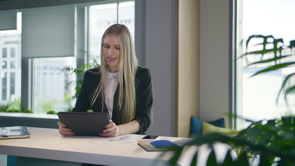 Elegant woman in suit sitting at table with laptop and surfing tablet in modern light office with big windows
