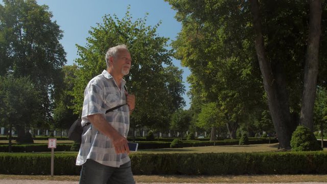 grey haired elderly man in checkered shirt walks along green park zone against clear blue sky close view slow motion. Concept historical heritage excursion