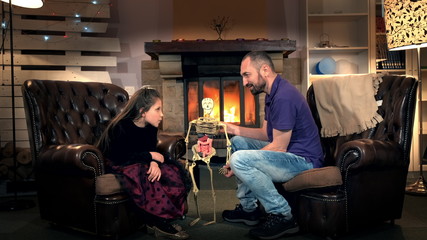 Naklejka na ściany i meble Little daughter giving father interesting questions about the human body structure. Dad holding in his hand human skeleton. In the background - fireplace.