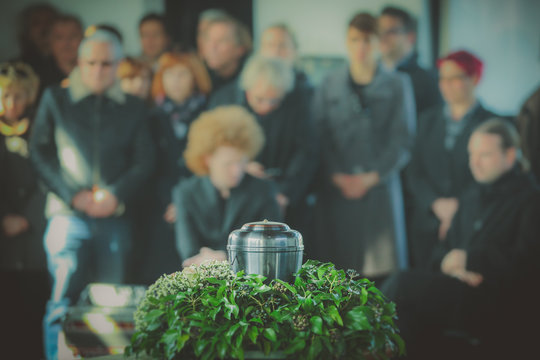 A metal urn with ashes of a dead person on a funeral, with people mourning in the background on a memorial service. Sad grieving moment at the end of a life. Last farewell to a person in an urn.