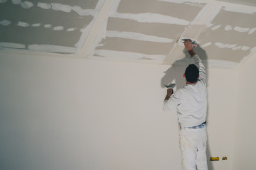 Man dressed in white, a painter, is applying putty or stucco to a dry wall installation in a house....