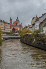Fototapeta na wymiar Picturesque buildings along Ljubljanica river on a dull autumn november day. Lazy gray day in Ljubljana, Slovenia, with dirty brown river flowing under the bridge.