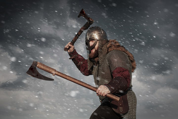 Warrior Viking in full arms with axe helmet and shield on dark sky background. Medieval scandinavian berserk red beard and fur costume attacks enemy. Concept historical photo