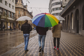 Three men walking with umbrellas, with a huge rainbow colored umbrella in between. Stroll with a rainbow umbrella in the centre of Ljubljana, Slovenia on a rainy day.