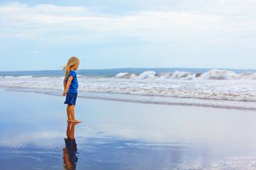 Child stand on black sand sea beach. Dreaming child look at sea surf landscape, breaking storm waves. Solitude concept. Retreat leisure on summer family vacation with kids. Travel lifestyle banner.