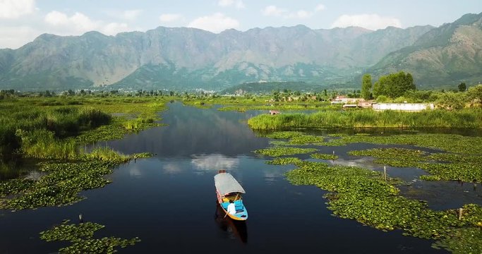 Panoramic aerial backward fly above Shikara floating on silent Dal Lake in Kashmir, India with mountainious landscape and blue sky in the background.
