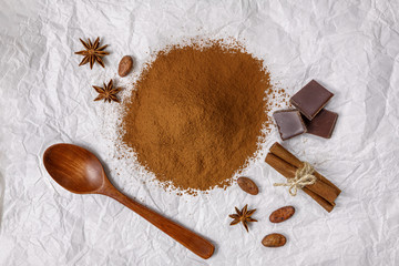 ground cocoa and cocoa beans with chocolate on a wooden spoon and cinnamon on crumpled white paper, top view