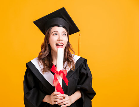 happy young woman in graduation gowns holding diploma and looking away