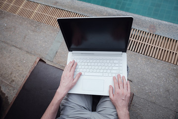 travel blogger sitting at swiming pool writing article on white laptop.