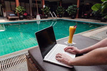 travel blogger sitting at swiming pool writing article on white laptop.
