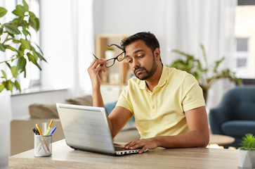 remote job, technology and people concept - bored or tired young indian man in glasses with laptop...