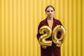 Young beautiful sad girl in burgundy coat and mustard sweater holds in her hands the helium balloons of number 20 against the background of bright yellow wall. Birthday concept in 20 years.