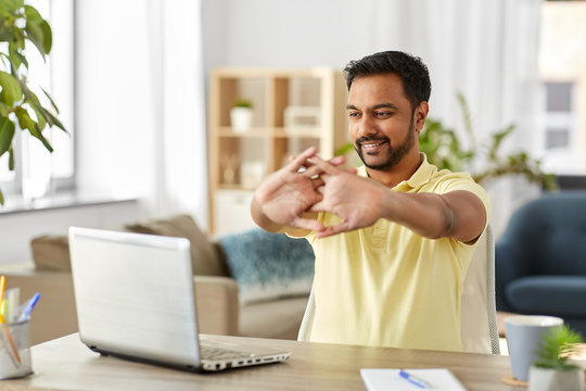 Technology, Remote Job And Lifestyle Concept - Happy Smiling Indian Man With Laptop Computer Stretching Arms At Home Office