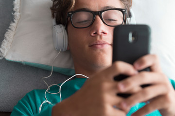 Young handsome caucasian teenager use cellular phone device and listen music with headphones lay...