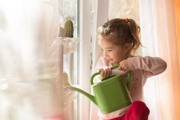 A little girl with a green watering can is playing on the window of the house and watering the...