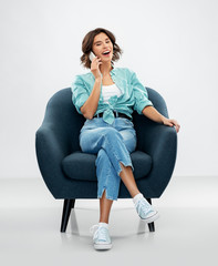 people and technology concept - portrait of smiling young woman in turquoise shirt and jeans sitting in modern armchair and calling on smartphone over grey background