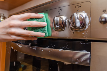 woman's hand washes a green sponge with foam on the switch handle of a gray stainless steel gas stove. Cleaning the apartment