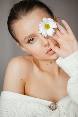 Obraz na płótnie Canvas Lovely girl with closed eyes in a white sweater brings a white flower to her eye and looks at the camera