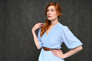 Portrait of a young pretty red-haired woman in a blue dress on a gray background. The model shows different emotions.
